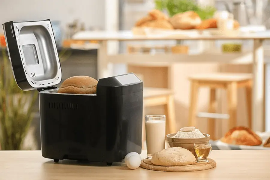 Máquina para hacer pan sobre una mesa con algunos panes y leche en una cocina casera