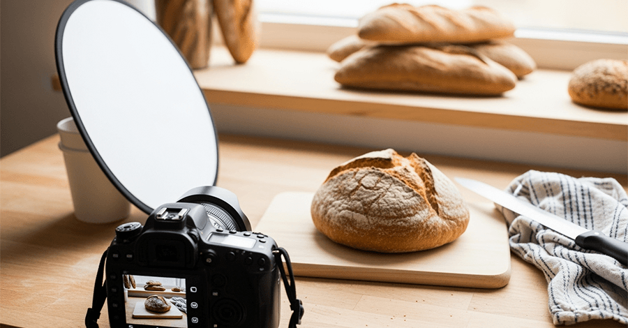 Cámara fotografiando pan rústico en una panadería artesanal, con reflector casero y utensilios simples sobre mesa iluminada por luz natural.