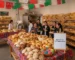 Panadería festiva con papel picado y manteles de colores; mesa frontal con conchas, bolillos y pan de fiesta junto a cartel “Feria del Pan y la Gastronomía”; gente degustando y horno/amasadora al fondo.