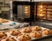 Obrador profesional en producción de Pan de Muerto: en primer plano, charola con piezas doradas y uniformes con huesitos y bolita bien formados; al fondo, horno industrial de convección encendido con el interior iluminado y más charolas horneándose, transmitiendo profesionalismo, control técnico y calidad consistente.