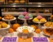 Vitrina frontal impecable de panadería moderna decorada para octubre, con Pan de Muerto, conchas y galletas temáticas exhibidas a diferentes alturas; flores de cempasúchil, calabazas pequeñas y papel picado naranja y morado enmarcan la escena. Iluminación cálida que realza dorados y texturas, creando un ambiente acogedor y festivo sin saturar la composición.