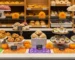 Vitrina frontal impecable de panadería moderna decorada para octubre, con Pan de Muerto, conchas y galletas temáticas exhibidas a diferentes alturas; cempasúchil, calabazas pequeñas y papel picado naranja y morado enmarcan la escena. Iluminación cálida que realza dorados y texturas, creando un ambiente acogedor y festivo sin saturar la composición.