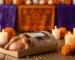 Altar de Día de Muertos en panadería artesanal con Pan de Muerto en empaque temático elegante sobre mesa de madera rústica, rodeado de cempasúchil y velas encendidas; al fondo, papel picado morado y naranja. Iluminación cálida que resalta la textura y el azúcar del pan, comunicando tradición y valor cultural con presentación premium.
