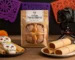 Mesa de madera rústica con cempasúchil, velas y papel picado naranja/morado; en el centro, Pan de Muerto en empaque elegante, acompañado de galletas de calaverita y tamalitos dulces. Al fondo, cámara enfocando la escena y smartphone con una publicación de Instagram. Iluminación cálida que resalta texturas y comunica tradición + estrategia visual para ventas.